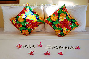 close-up of King bed with colourful pillows and welcome flowers spelling kia orana